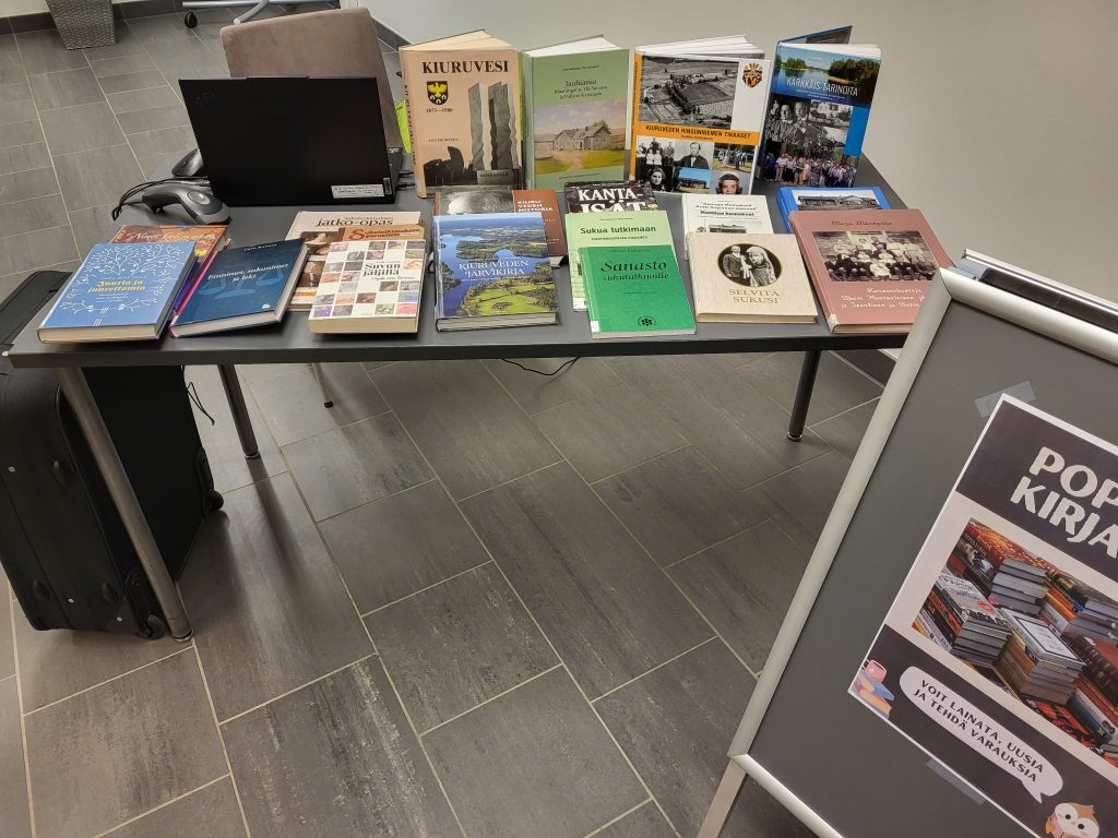 Kuvassa pöytä, jonka päällä on kirjaston kirjoja. Kuvassa näkyy myös matkalaukku ja ständi, jossa on pop up -kirjaston mainos.